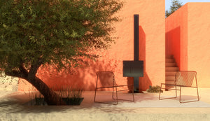 Der schwarze Gartenkamin von dyook steht in einem mediterranen Umfeld mit Olivenbaum und zwei Stühlen. Der moderne und gradlinige Gartenofen ist geschlossen und brennt nicht.